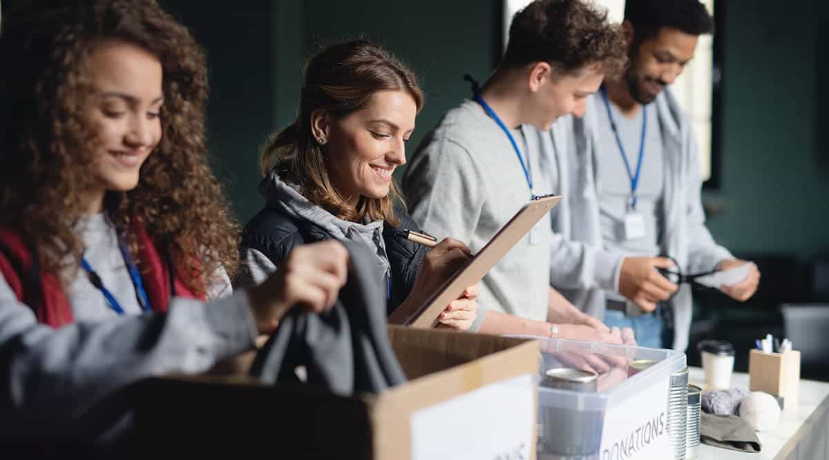 Four volunteers sorting donations into boxes at a table, one writing on a clipboard.