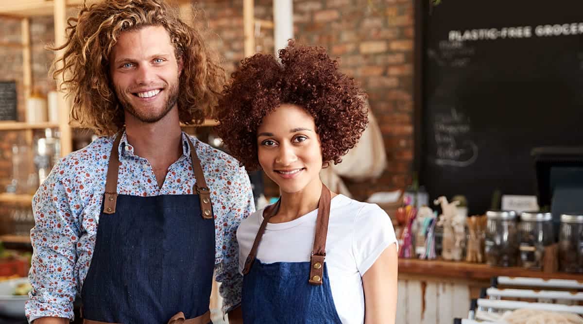 Two people in aprons standing in a café with shelves and jars behind them.