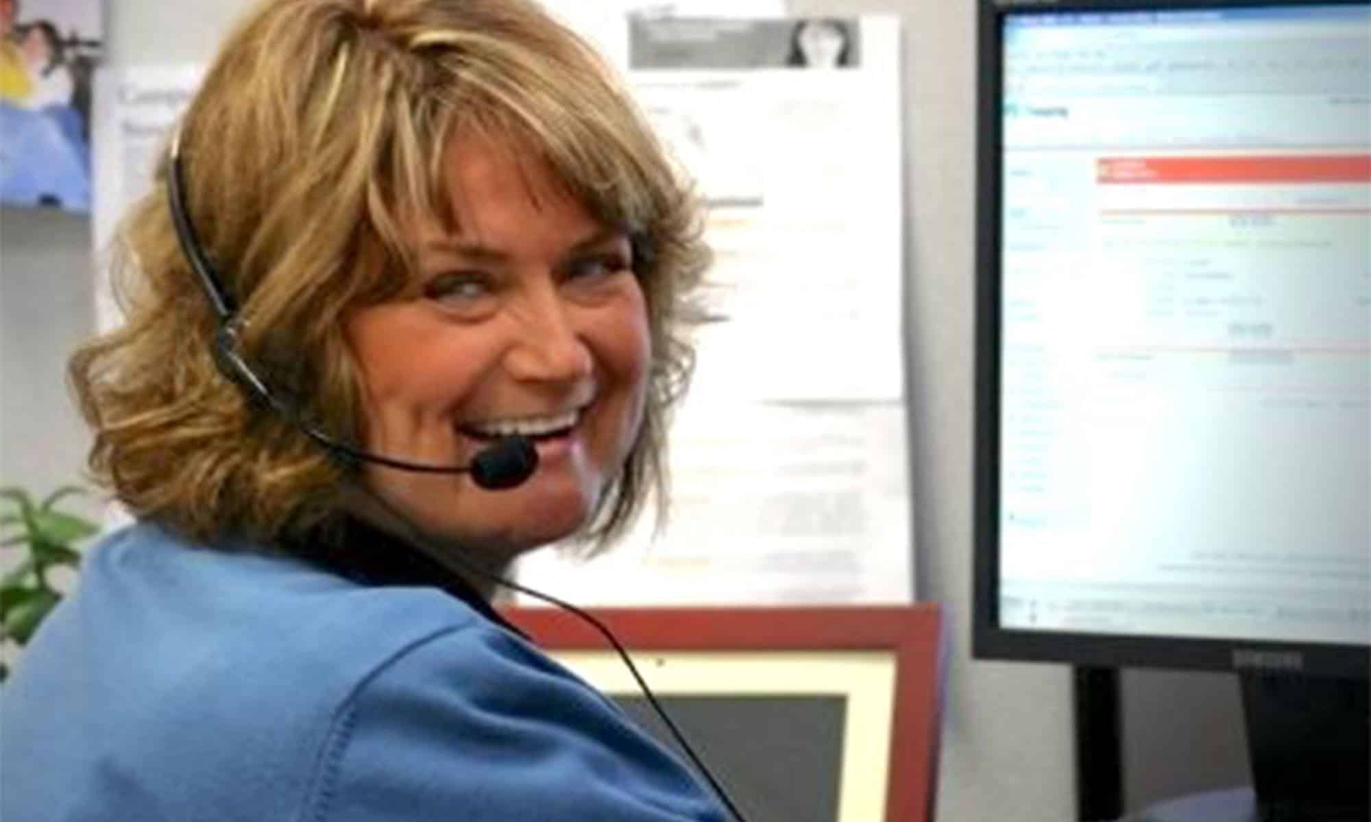 Woman with headset smiling at a desk with a computer monitor.