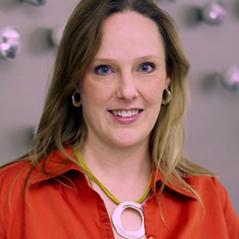 Jennie Martin A woman with light brown hair wearing a red shirt, hoop earrings, and a large geometric necklace stands in front of a blurred background.
