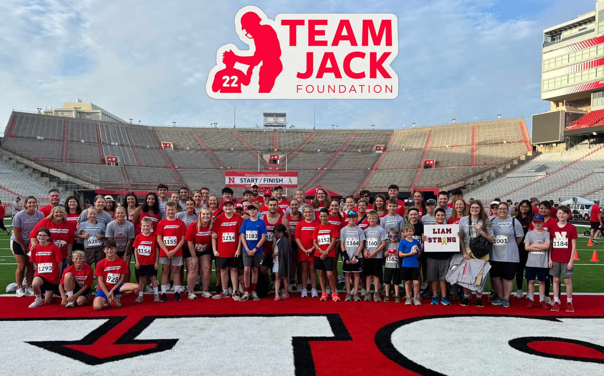 A large group stands on a football field holding race bibs and signs, under a “Team Jack Foundation” banner, preparing for an athletic event.